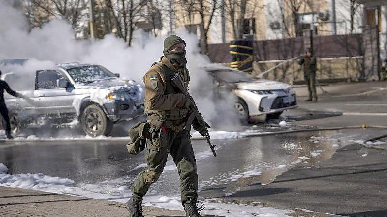 Ein ukrainischer Soldat bezieht Stellung vor einer Militäreinrichtung, während Feuerwehrleute im Hintergrund zwei brennende Autos löschen. Foto: Emilio Morenatti/AP/dpa